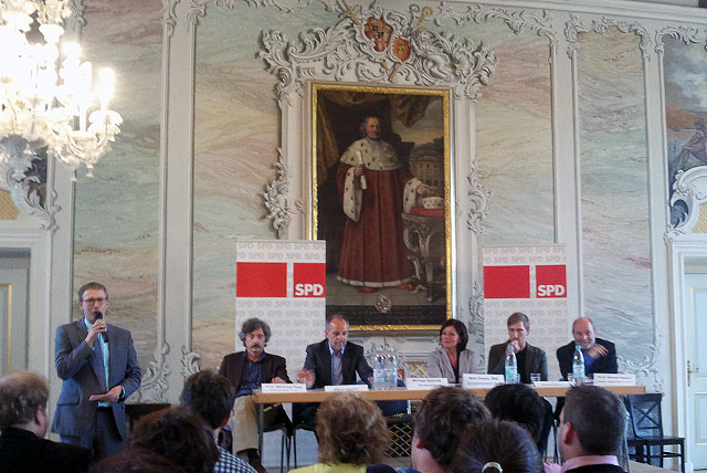 Menschen sitzen auf einem Podium, im Hintergrund SPD-Schilder und ein barockes Gemälde